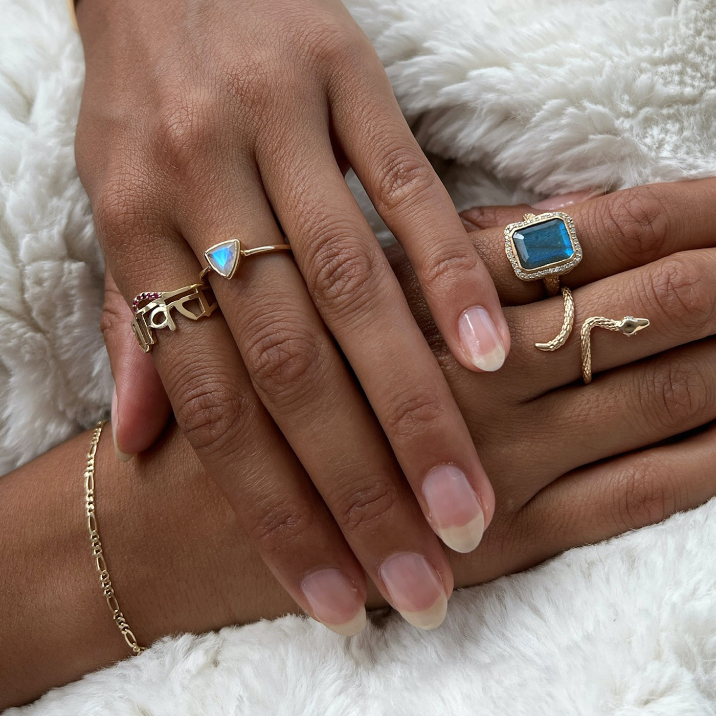 Hand model wearing multiple yellow gold rings. The first is a Sanskrit ring with rubies, the second is a trillion cut moonstone, the third is a emerald cut labradorite and the last is a textured gold snake ring.