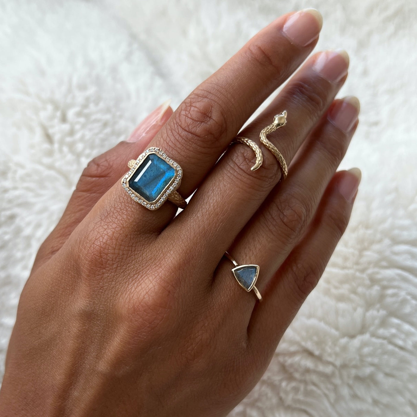 Hand model wearing three rings. The first ring is a emerald cut labradorite with a diamond halo, the second ring is a textured gold snake ring, the third ring is a trillion cut labradorite.