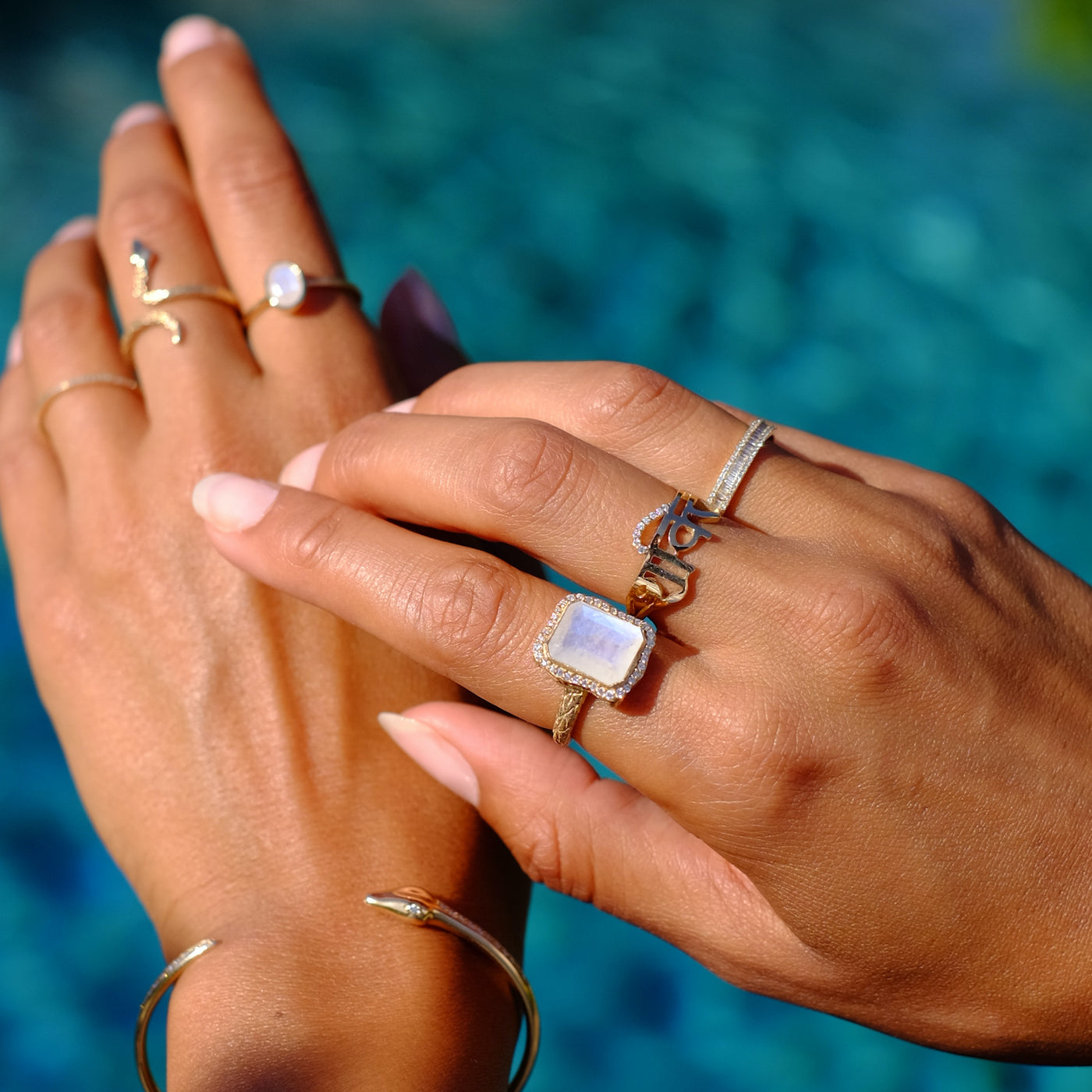 Hand model wearing six yellow gold rings. The ring on her pointer finger is an emerald cut moonstone on a textured gold band with a diamond halo.