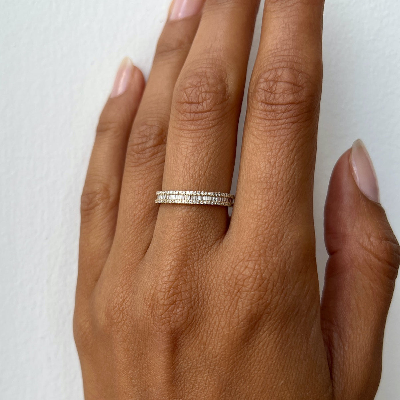 Hand model wearing a yellow gold diamond band which features two bordering rows of round diamonds with a center row of tapered baguette diamonds.