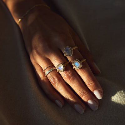 Hand model wearing three yellow gold rings with moonstones. Each ring features a different shape stone. Pictured are trillion cut, emerald cut and pear cut moonstones.